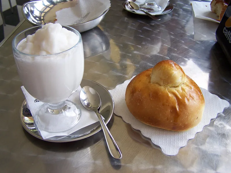 A glass of almond granita next to a round brioche col tuppo at a Sicilian café